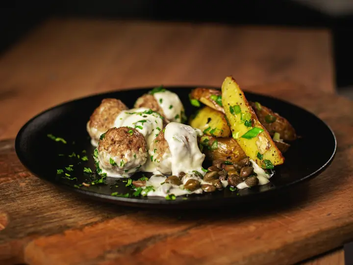 Frikadeller i cremet sauce anrettet på en sort tallerken sammen med ovnbagte kartoffelbåde