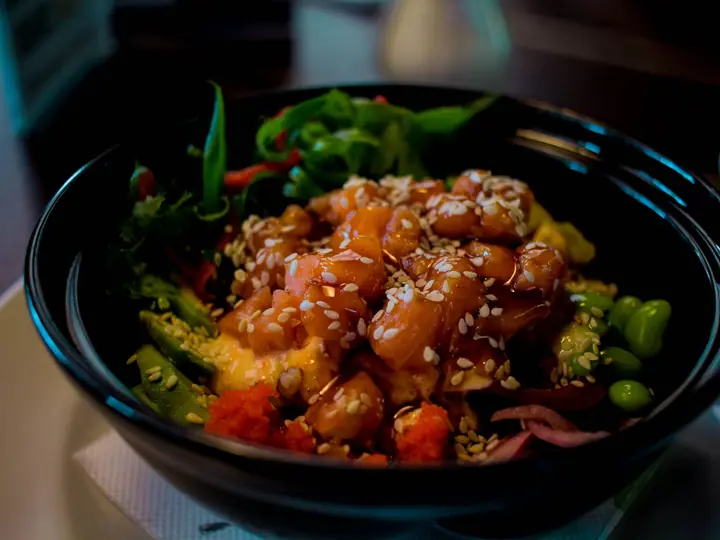 Sort skål med terninger af laks i blank dressing, drysset med sesam, med edamamebønner, avocado, rødløg og grønne blade langs kanten